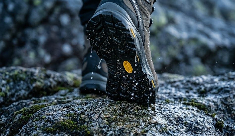 Podeszwa buta trekkingowego z agresywnym bieżnikiem i widocznym żółtym logo Vibram 2026, stojąca na mokrej, mszystej skale w górach. Podeszwa buta trekkingowego z agresywnym bieżnikiem i widocznym żółtym logo Vibram 2026, stojąca na mokrej, mszystej skale w górach.